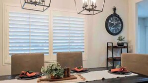 A dining area with a long table, two chairs, a clock on the wall, and a window with blinds.