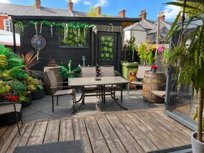 A wooden deck with a table and chairs, surrounded by potted plants and a black wall with a clock.