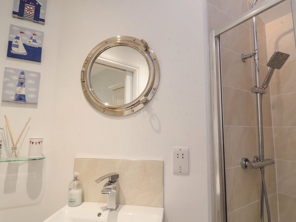 Un baño con un espejo redondo, un lavabo con grifo moderno, un estante de vidrio con artículos de aseo y una zona de ducha con puerta de cristal.