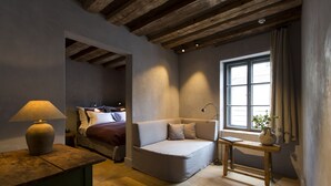 Una habitación acogedora con techo de madera, una cama con manta roja, un sofá color beige, una mesita con lámpara y una ventana con cortinas.