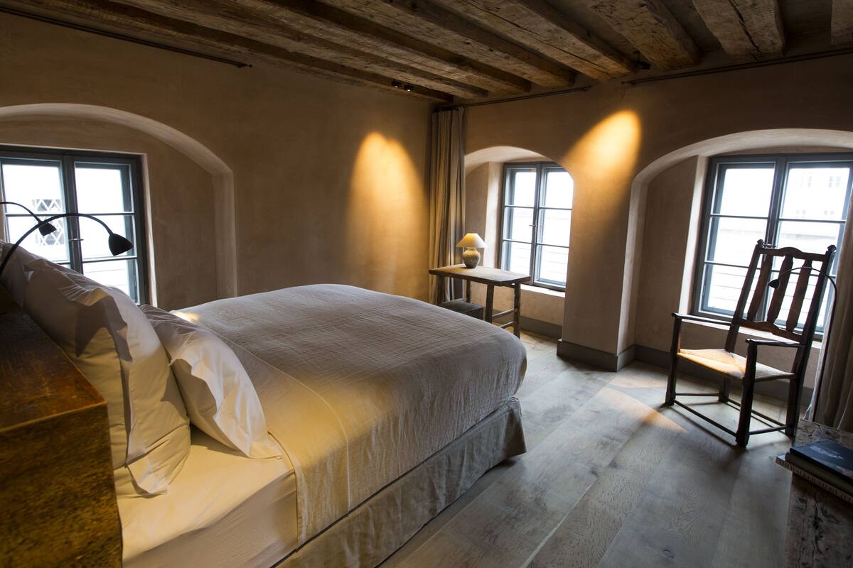 Un dormitorio con cama, una silla, una mesa y dos ventanas.