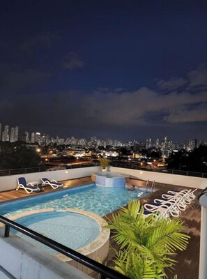 Una piscina en la azotea con sillas de descanso y un jacuzzi, con vistas a un paisaje urbano nocturno.