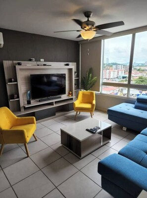 Una sala de estar moderna con un televisor de pantalla plana, sillas amarillas y azules, una mesa de centro y un ventanal con vista a la ciudad.