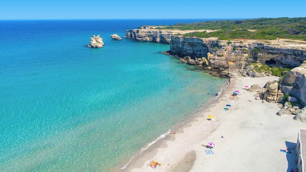 Uma praia costeira com águas turquesa cristalinas, falésias rochosas e uma faixa de areia com guarda-sóis e pessoas.