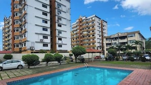A rectangular swimming pool with a brick border, surrounded by multi-story residential buildings with balconies, under a clear blue sky.