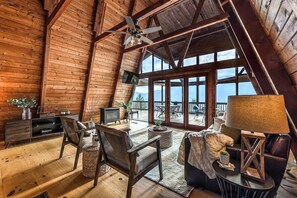 A cozy wooden interior with a high ceiling, a fireplace, comfortable seating, and large windows offering a view.