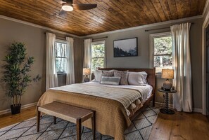 A bedroom with a wooden ceiling, a large bed with a headboard, two windows with curtains, a bedside table with a lamp, and a potted plant.