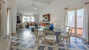 Un coin repas avec une table en verre, des chaises blanches et un vase bleu servant de centre de table. On aperçoit un canapé avec des coussins orange à l’arrière-plan, ainsi qu’une fenêtre avec des rideaux.
