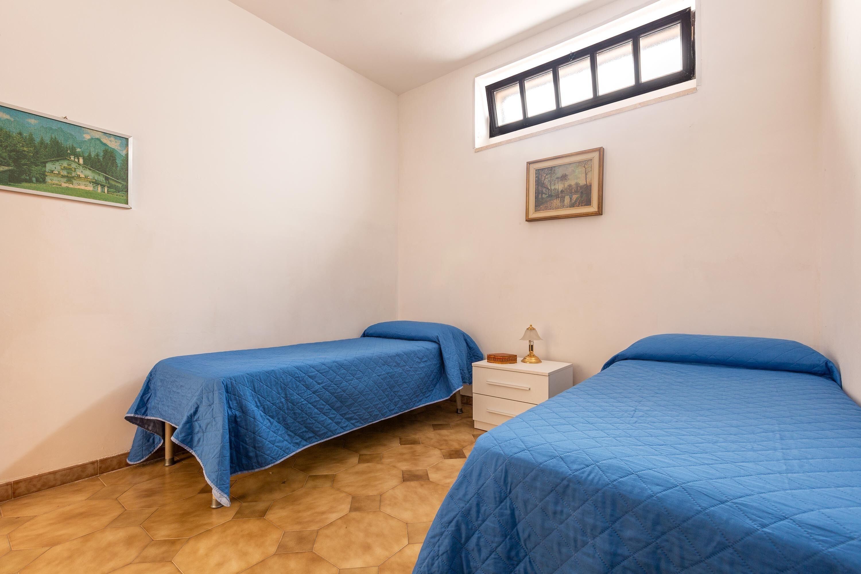 A bedroom with two beds, a small bedside table, a window, and a painting on the wall.