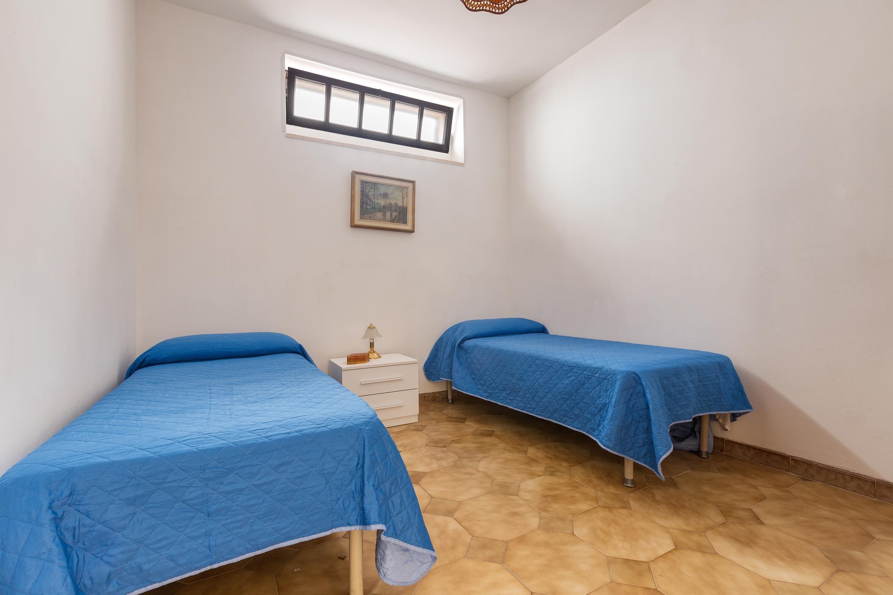Quarto pequeno com duas camas, uma janela, mesa de cabeceira e um quadro na parede.