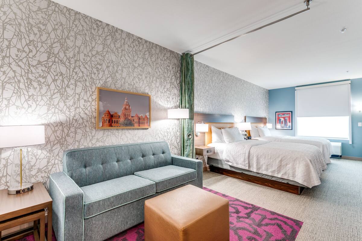 Een hotelkamer met een grijze bank, een bed met wit beddengoed, een nachtkastje met een lamp en een lijst met een foto aan de muur.