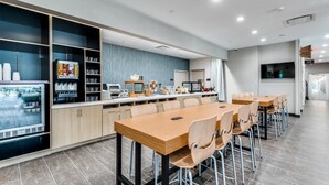 A modern dining area with a long wooden table and multiple chairs, a refrigerator with beverages, and a counter with various food items and a coffee machine.