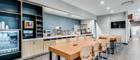 Uma sala de jantar moderna com uma mesa de madeira comprida e várias cadeiras, um refrigerador com bebidas e um balcão com diversos alimentos e uma máquina de café.