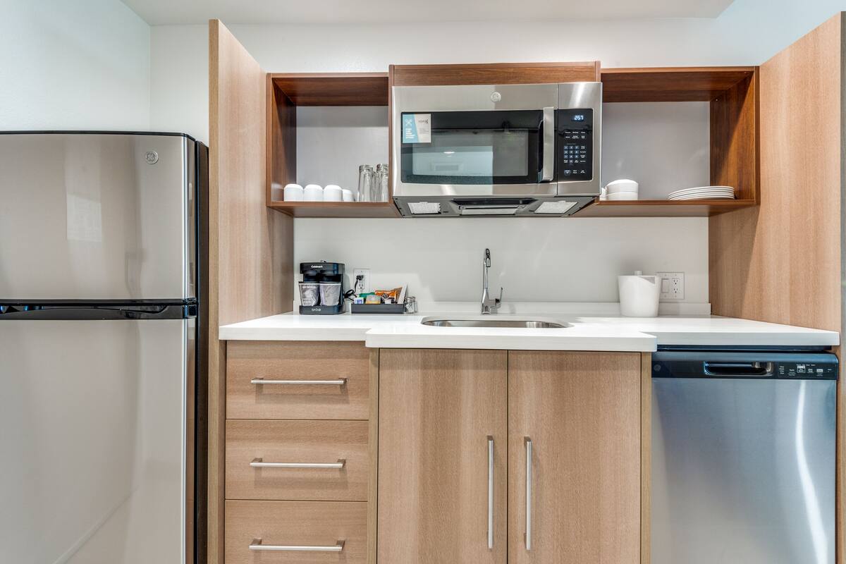 Een moderne keuken met een roestvrijstalen koelkast, magnetron en vaatwasser, houten kastjes en een wit aanrecht.