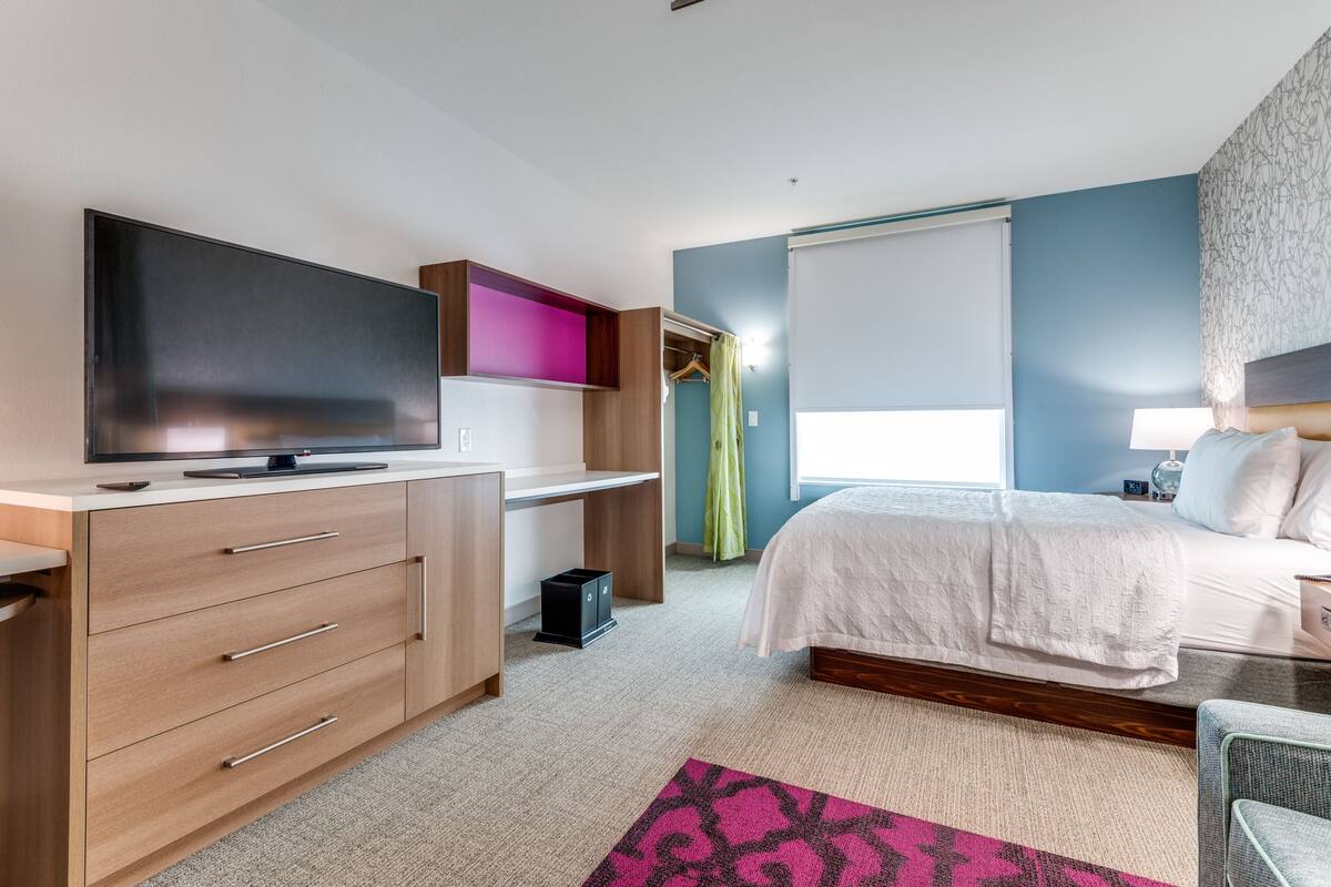 Een slaapkamer met een bed, een bureau met een flatscreen televisie, een kledingkast en een raam met gordijnen.