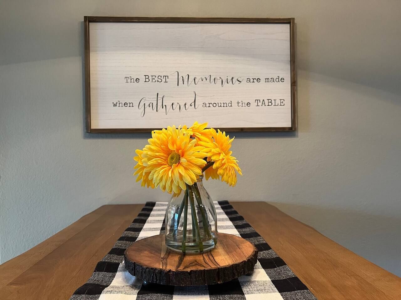 Uma placa emoldurada com texto, um vaso com flores amarelas e uma mesa com toalha xadrez.