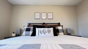 Une chambre à coucher bien rangée, avec un grand lit, une table de chevet avec une lampe et trois tableaux encadrés accrochés au mur.