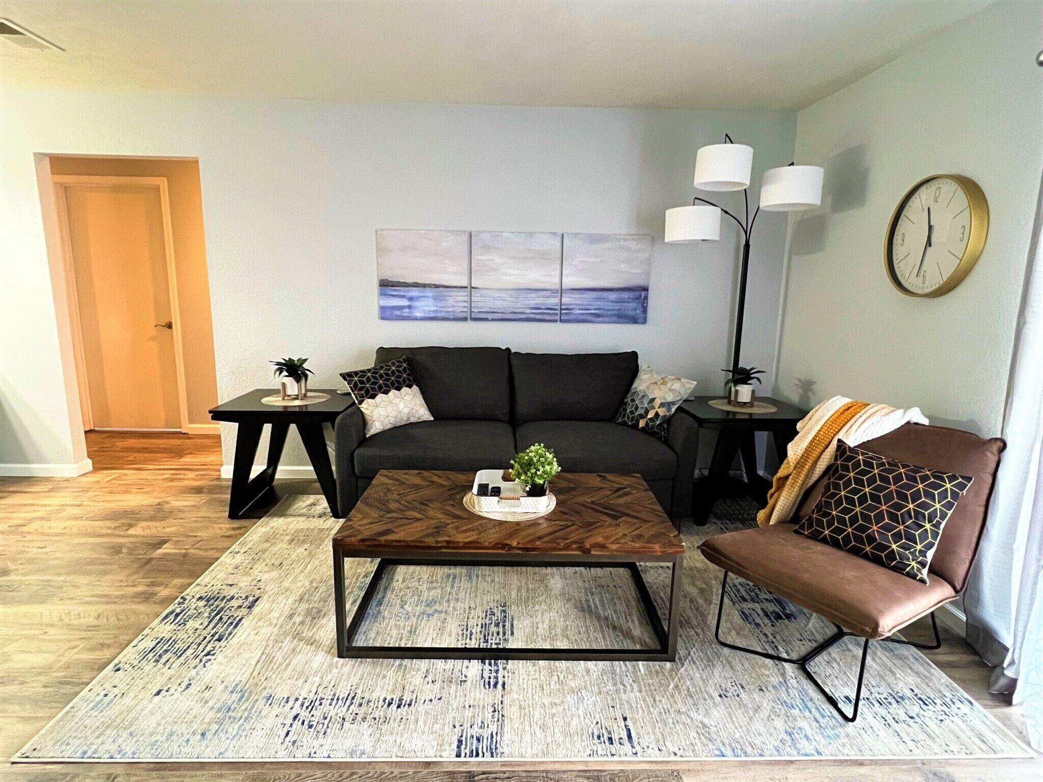 Een moderne woonkamer met een bank, salontafel en bijzettafeltjes. Aan de muur hangt een schilderij en er staat een klok.