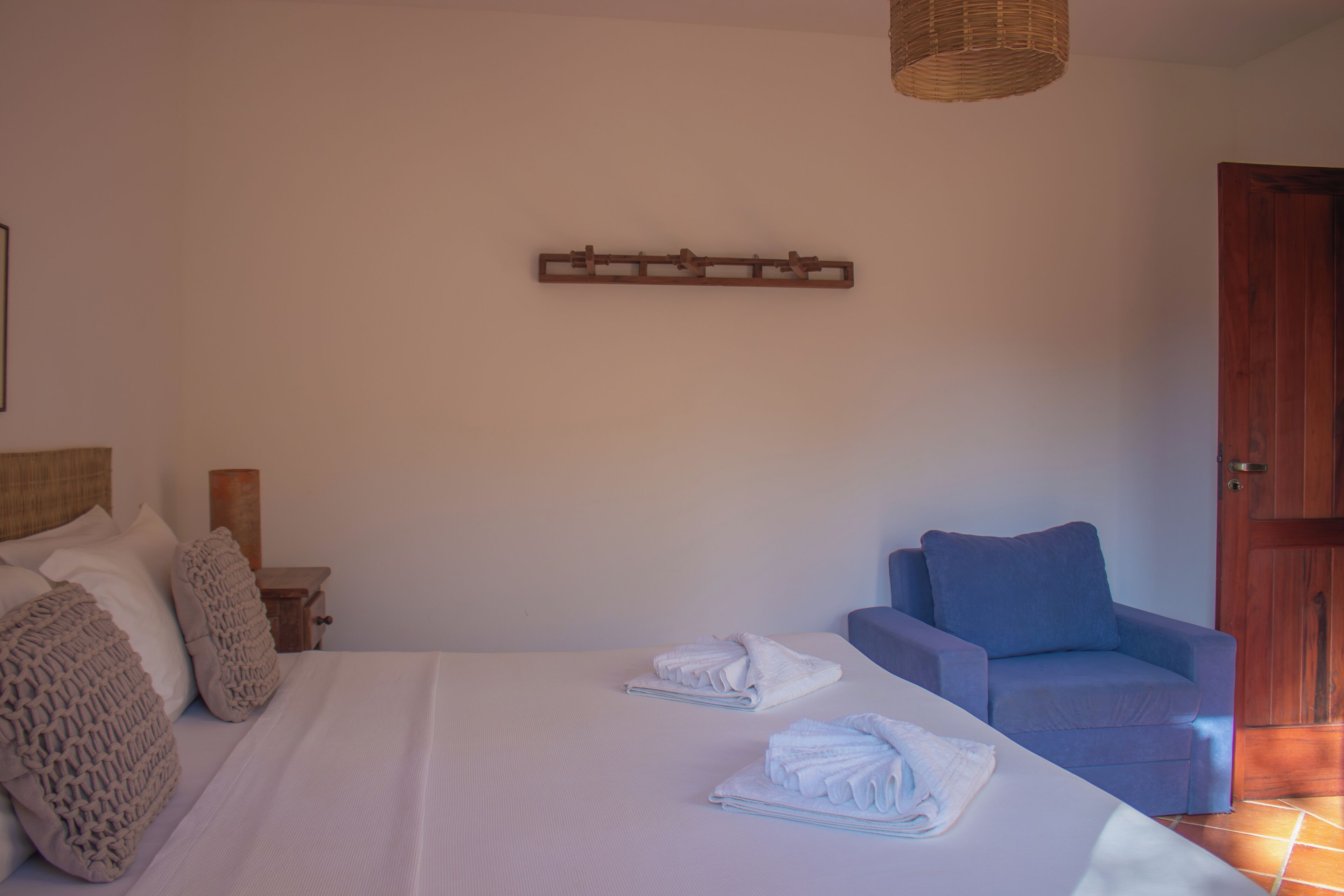 A bedroom with a bed, pillows, a chair, a wooden headboard, a wall-mounted shelf, and a door.