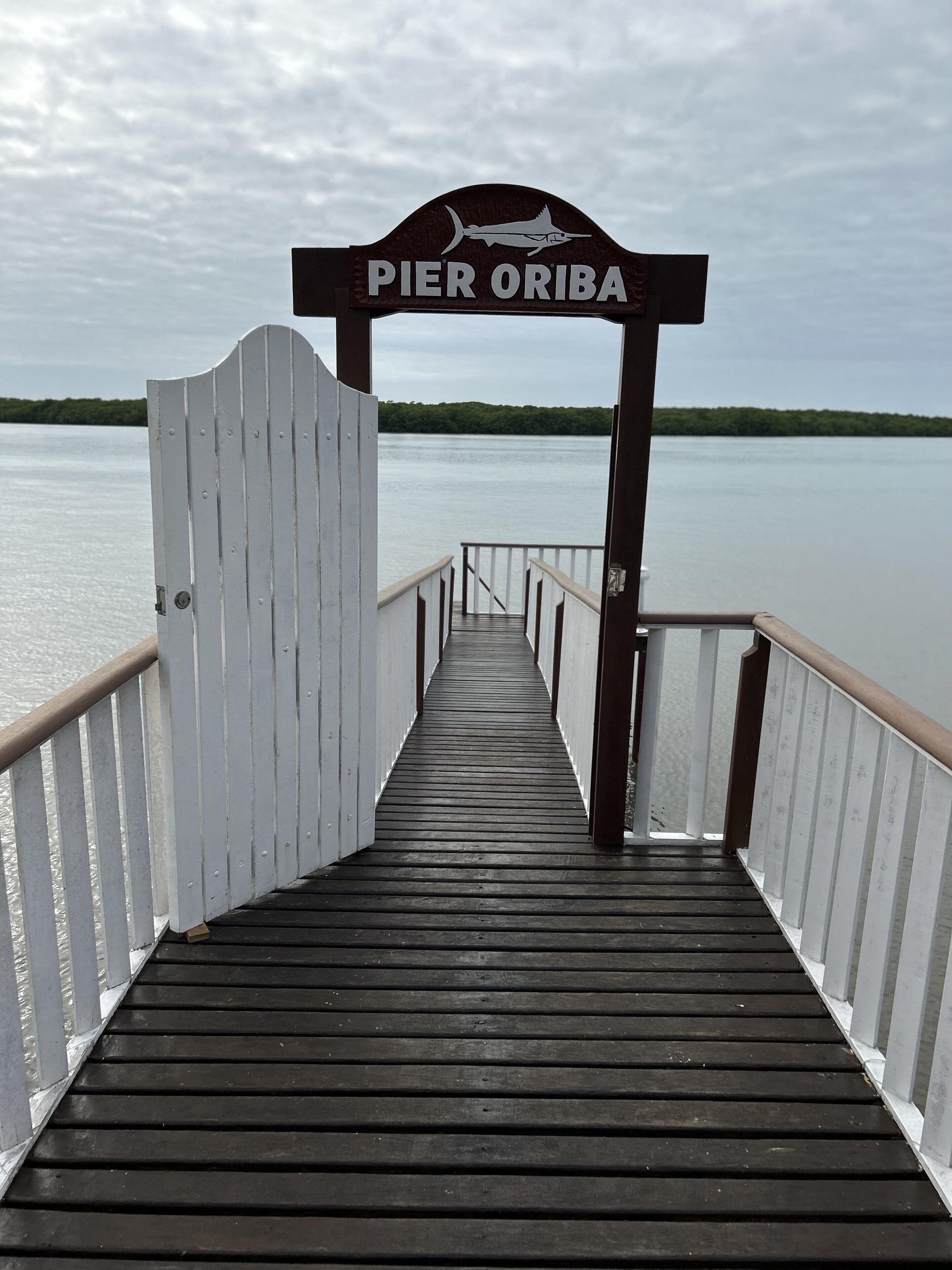 Un moll de fusta que condueix a Pier Oriba, amb vista a una aigua tranquil·la i una riba verda allunyada.