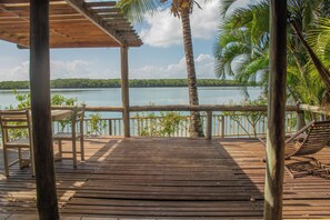A wooden deck with chairs and a table overlooking a lake, surrounded by palm trees. - Casa Oribà  (Santa Cruz Cabralia)