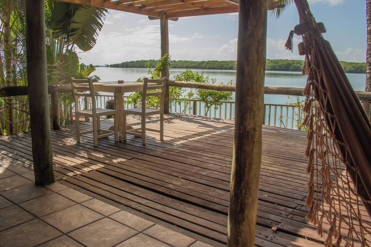 Una terraza de madera con una mesa y sillas, con vista a un lago y la vegetación circundante.