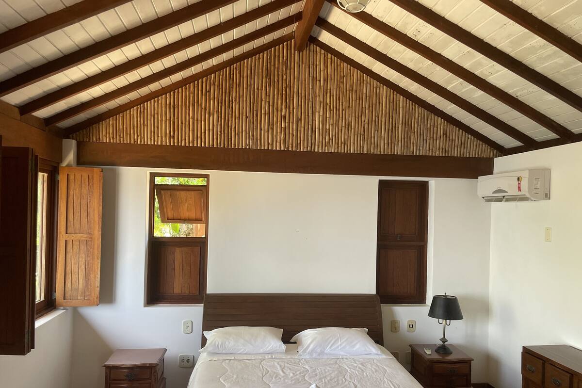 Un dormitorio con techo de madera, una cama con ropa de cama blanca, una mesita de noche, una lámpara y un aire acondicionado instalado en la pared.