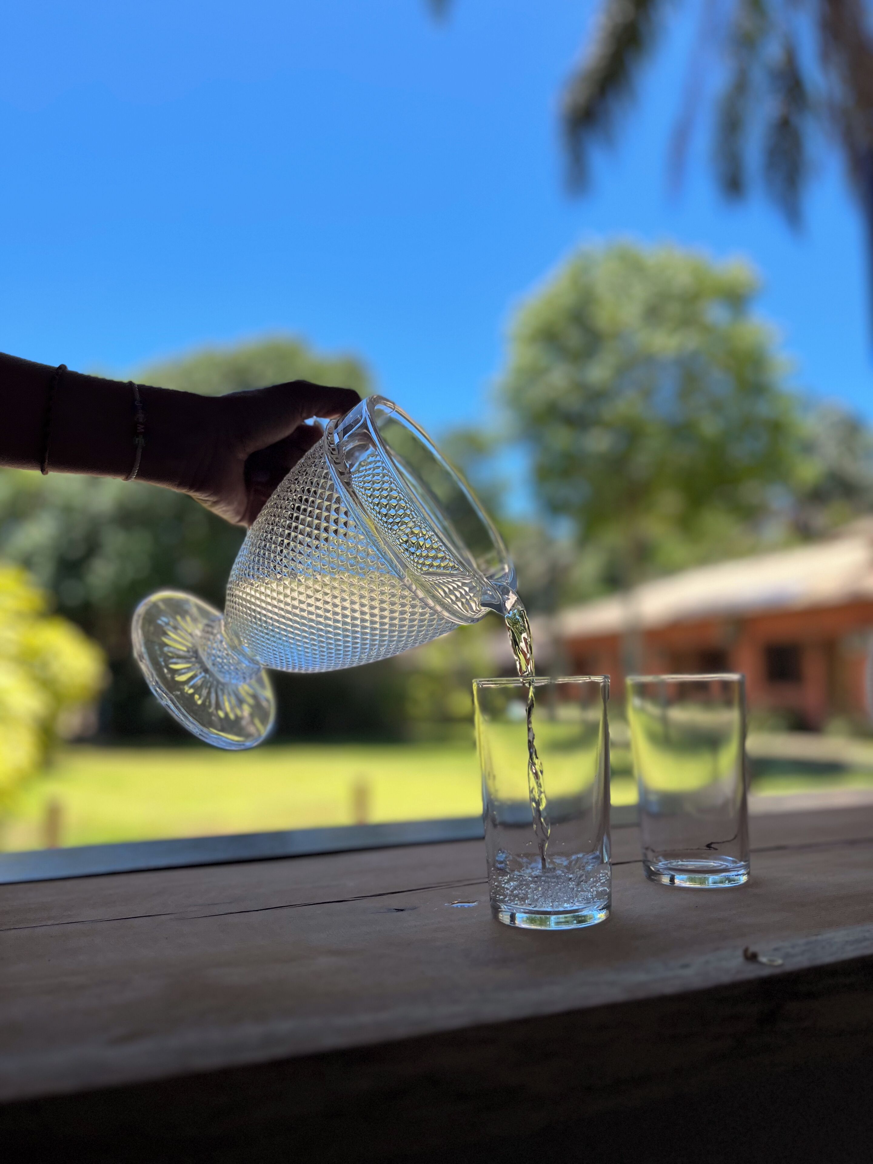 Una mano sirve agua de un jarro de vidrio transparente en dos vasos sobre una superficie de madera.