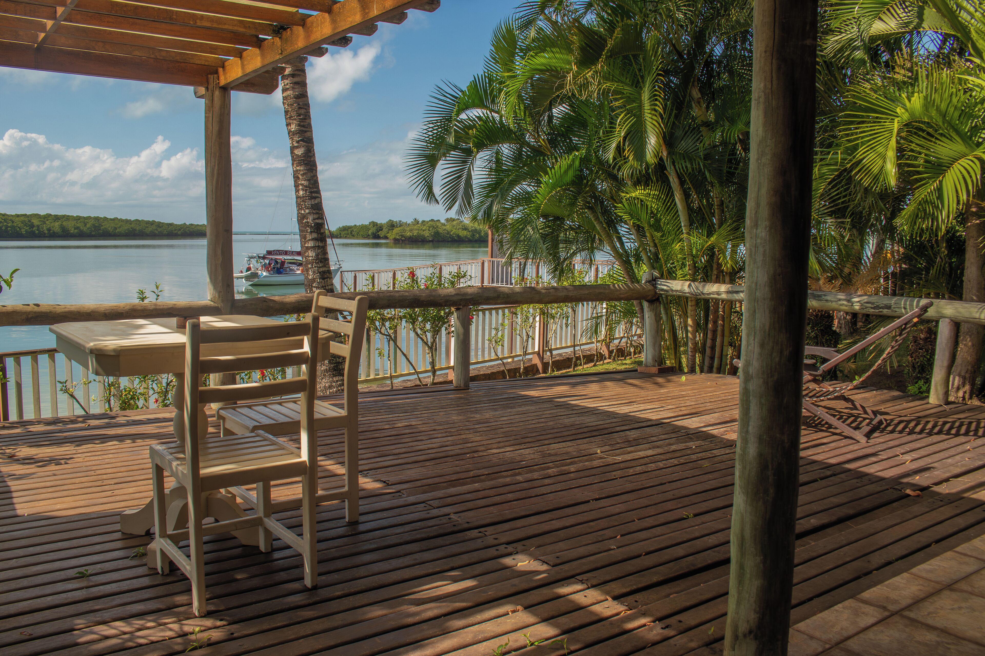 A wooden deck with a table and chairs, overlooking a calm lake with a boat in the distance.