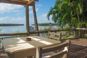 Una terraza de madera con una mesa y sillas con vistas a un lago tranquilo, rodeada de exuberante vegetación y un cielo despejado.