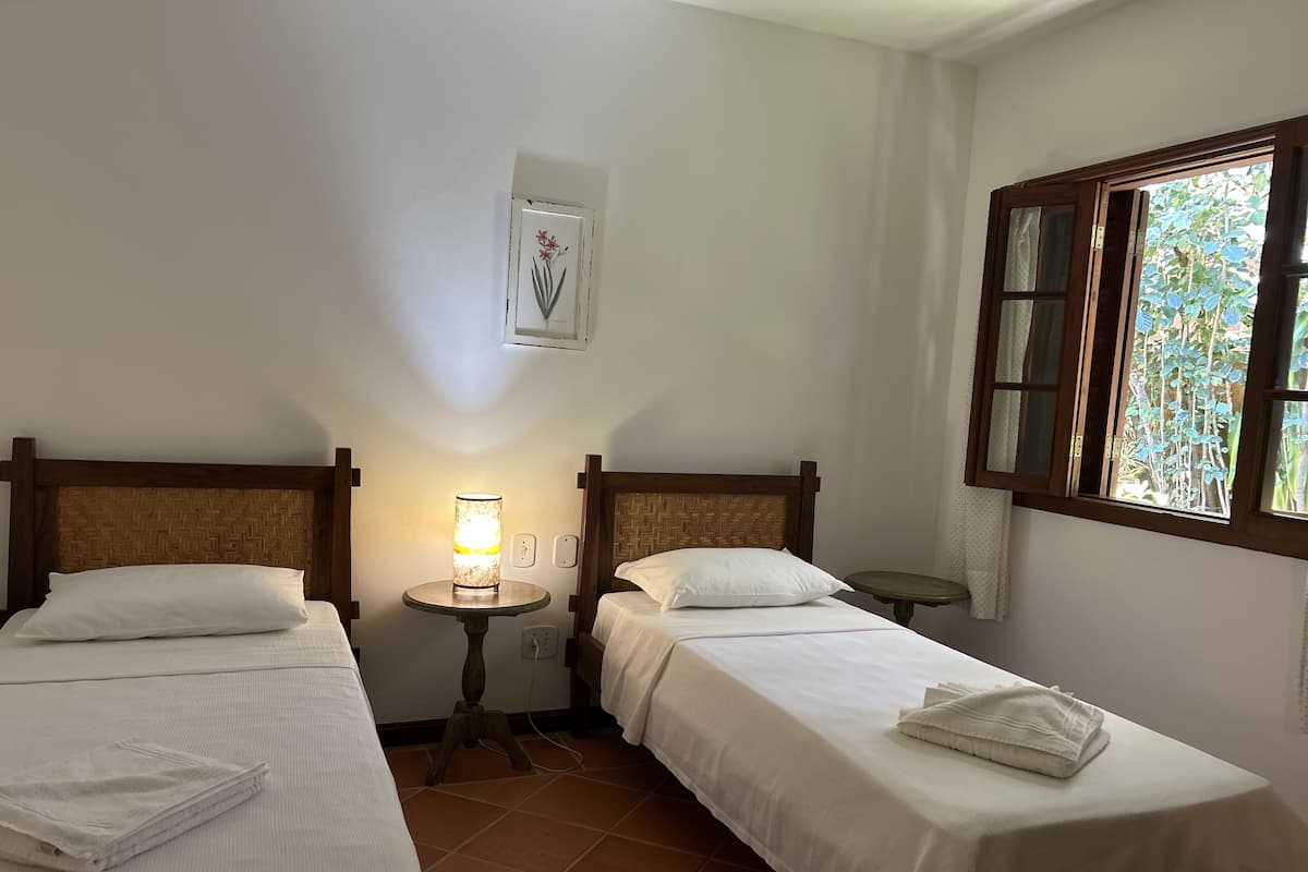 Habitación de hotel con dos camas individuales, una mesita de noche con lámpara, una pequeña mesa redonda y ventana con vistas a zonas verdes.