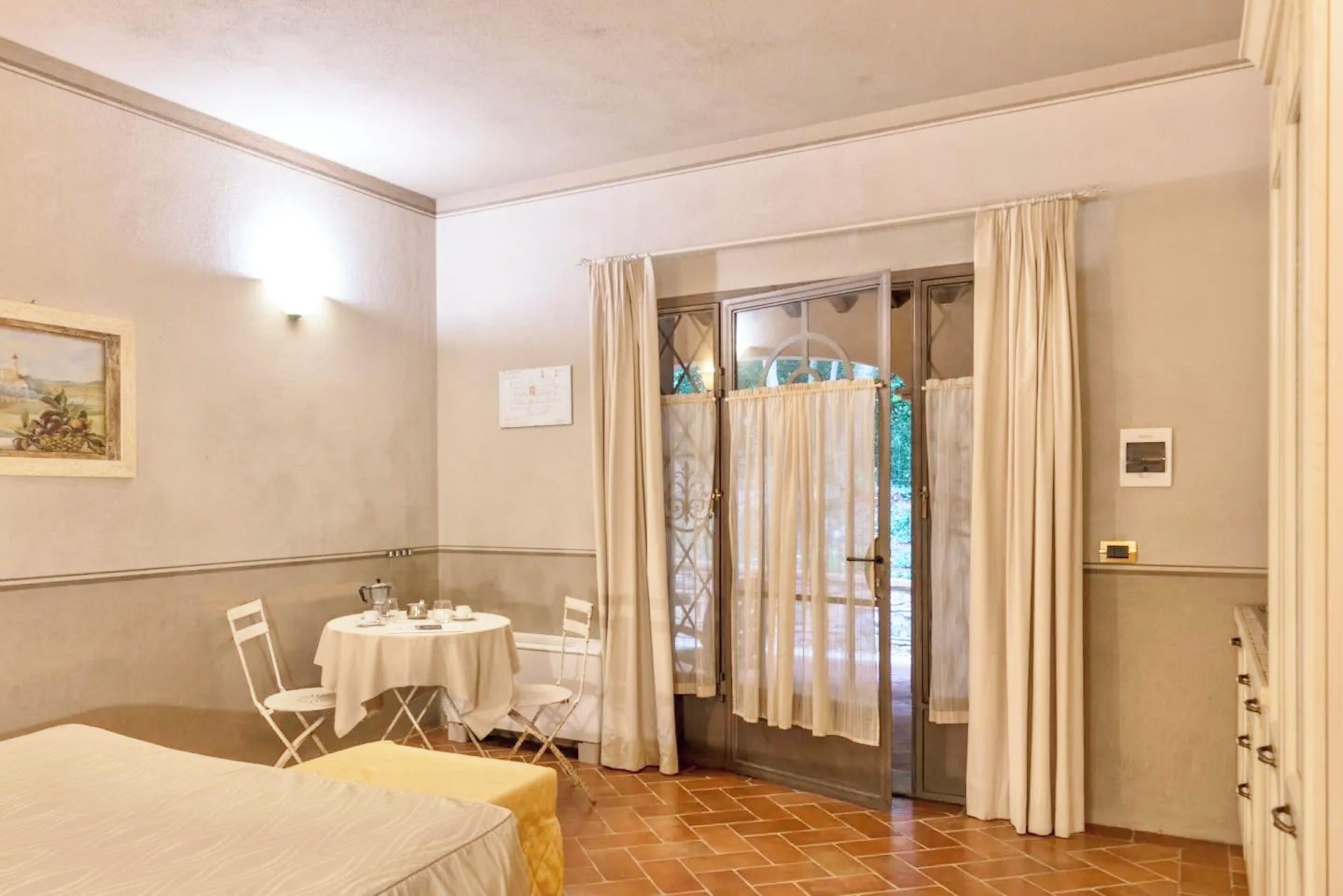 Una stanza con un letto, un tavolo e sedie, una finestra con tende e una porta che dà sull'esterno.