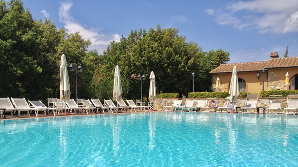 A clear blue pool with white sun loungers and umbrellas, surrounded by trees and a building with a tiled roof. - Residence con piscina a Monteverdi  (Monteverdi Marittimo)