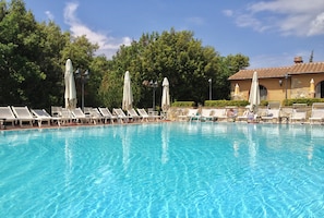 Une piscine d’un bleu éclatant, bordée de chaises longues et de parasols blancs, le tout entouré d’arbres et d’un bâtiment dont le toit est recouvert de tuiles.