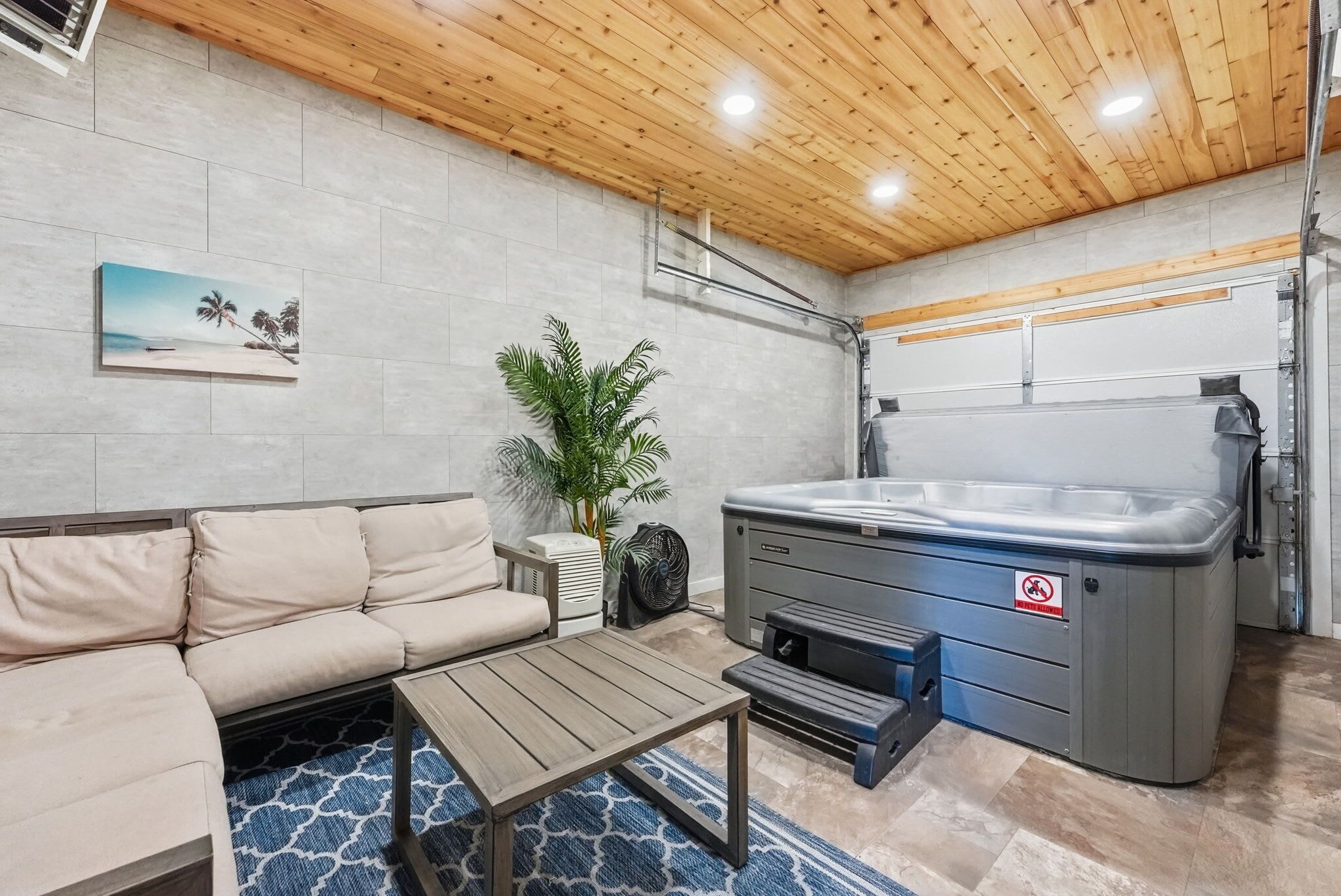 A room with a beige sofa, a small table, a hot tub, a plant, and a painting of a beach scene.