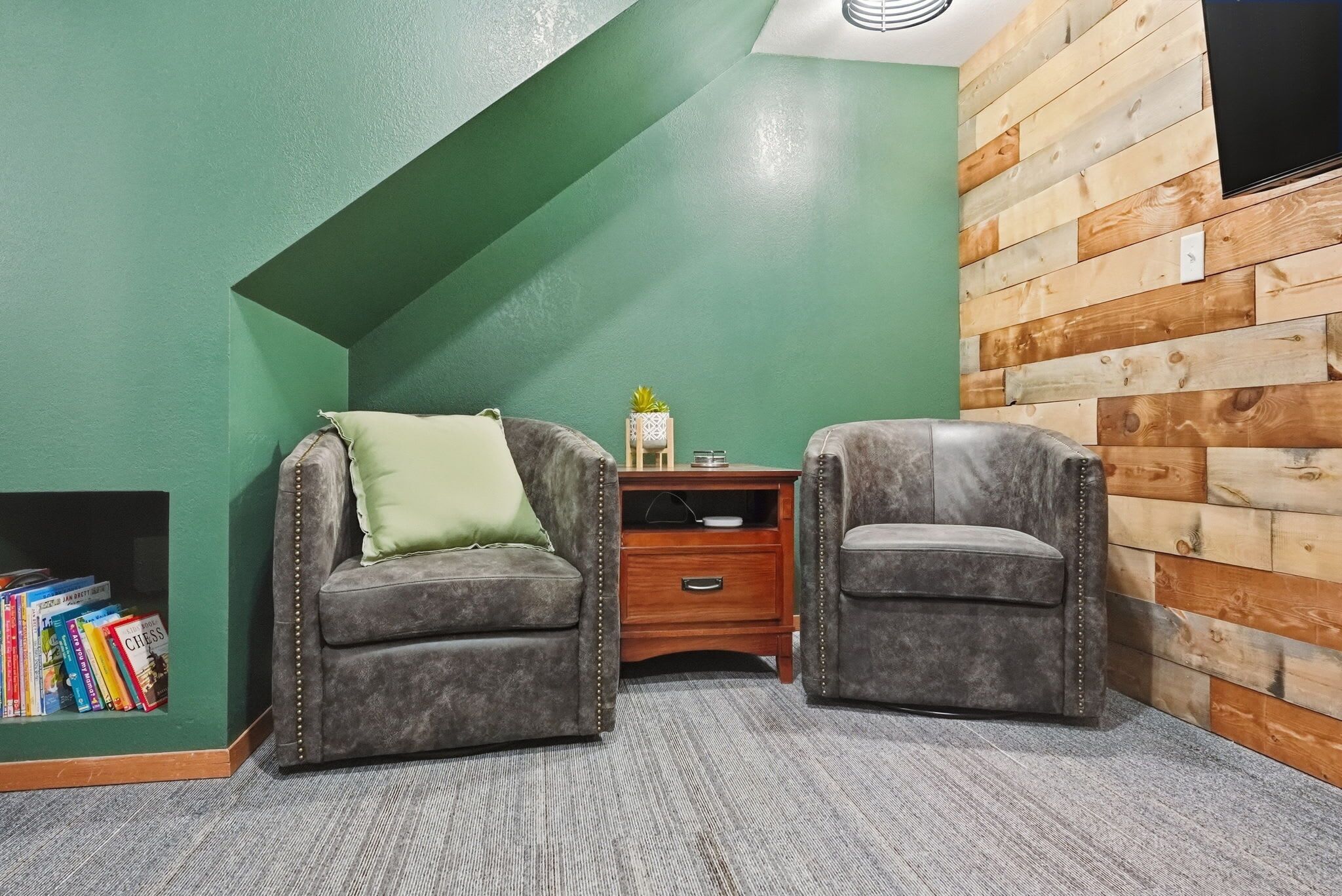 A room with two armchairs, a small wooden side table, and a bookshelf against a green wall.