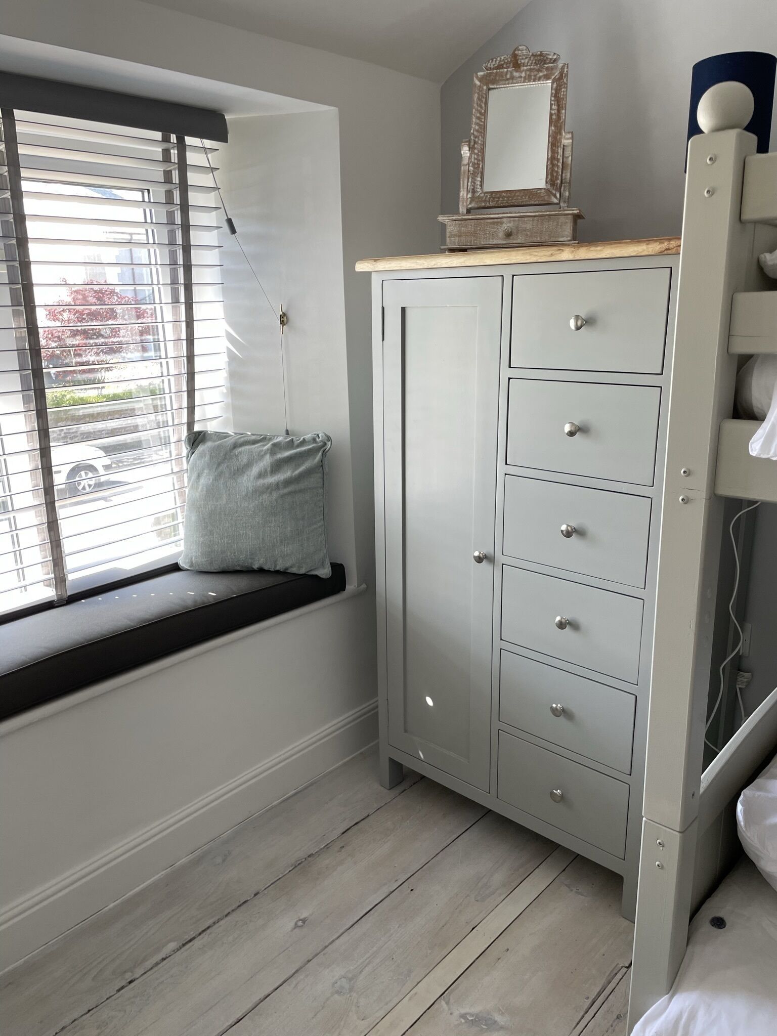 Una habitación con una ventana con asiento, un armario gris con varios cajones y un espejo encima.