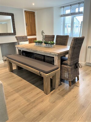 Un comedor con mesa de madera, sillas de mimbre, un banco y plantas en macetas.