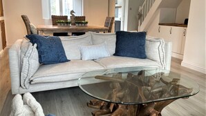 Una sala de estar con un sofá de color claro, dos cojines azules, una mesa de centro de vidrio con base de madera, sillas de mimbre y una zona de comedor con una mesa y sillas de madera.