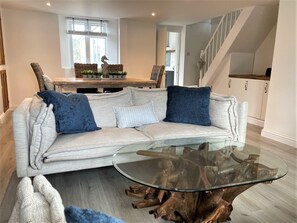 Un salon avec un canapé de couleur claire, deux coussins bleus, une table basse en verre avec un pied en bois, des fauteuils en osier et un coin repas avec une table et des chaises en bois.