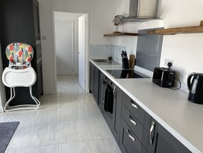 Une cuisine moderne avec des meubles de couleur sombre, un plan de travail blanc, un grille-pain, une bouilloire et une chaise haute colorée pour bébé.