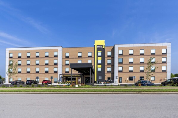 A modern hotel with a central entrance, multiple floors, and a parking lot with several cars. - Home2 Suites Lexington Keeneland Airport, KY (Lexington)