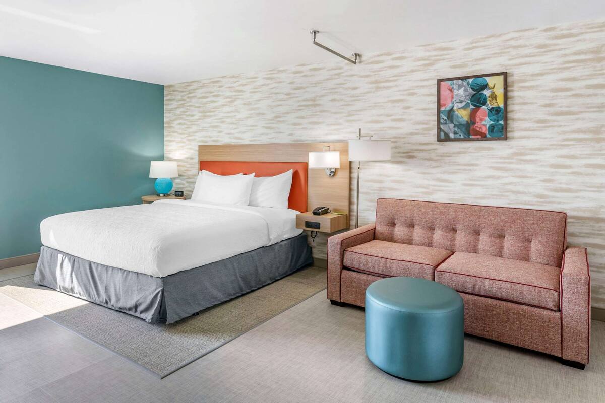 Une chambre d’hôtel moderne avec un grand lit, un canapé, un pouf rond et une œuvre d’art accrochée au mur.