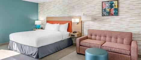 Una habitación de hotel moderna con una cama grande, un sofá, un otoman redonda y una obra de arte colgada en la pared.