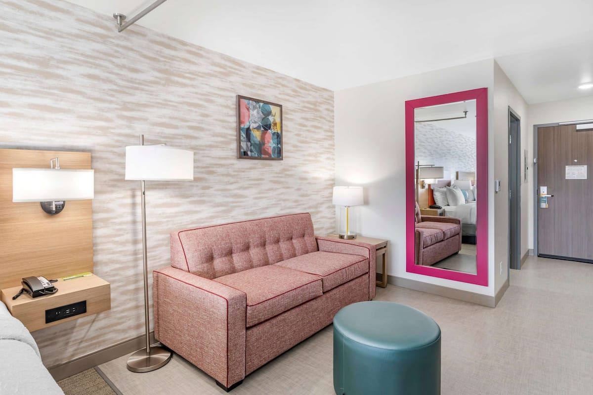 Une chambre d’hôtel moderne avec un canapé rose, un lampadaire, un miroir avec un cadre rose et un pouf bleu.