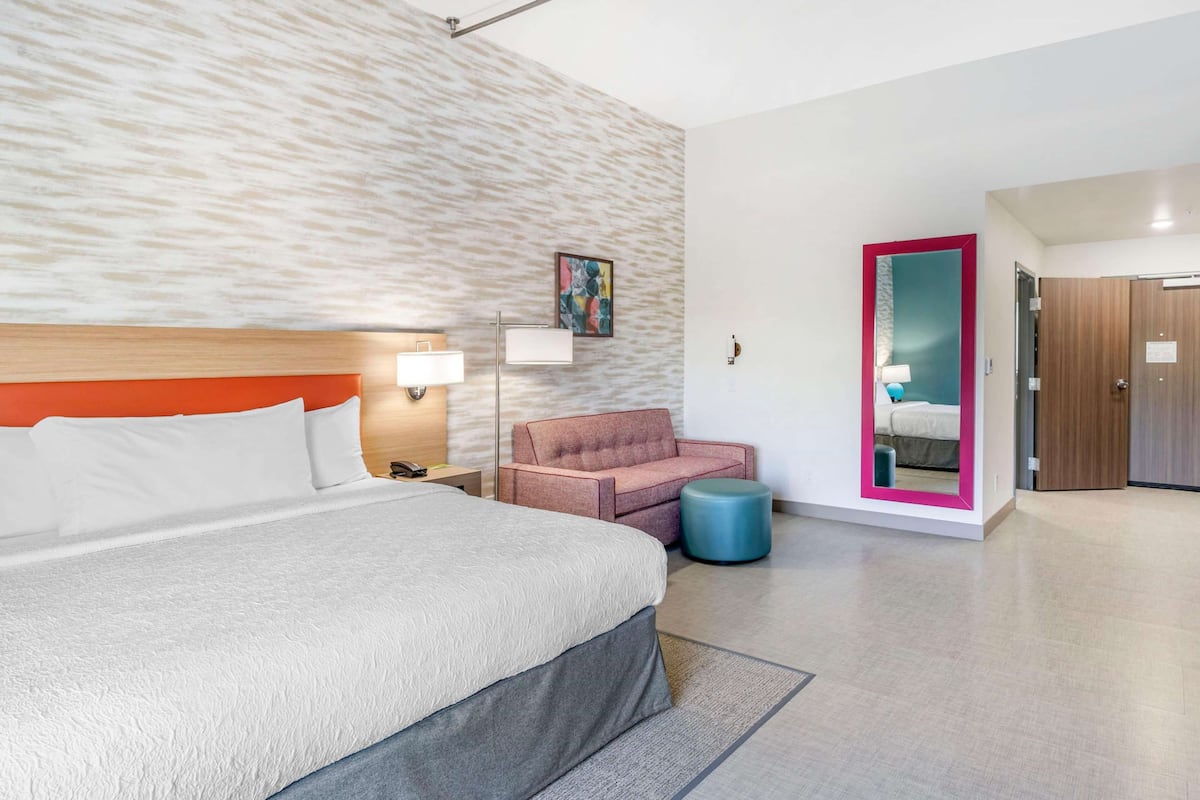 Une chambre d’hôtel moderne avec un grand lit, un fauteuil rose, un pouf rond, un miroir avec un cadre rose et une porte en bois.