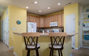 A kitchen with two wooden bar stools, wooden cabinets, a white refrigerator, a microwave, and a white door.