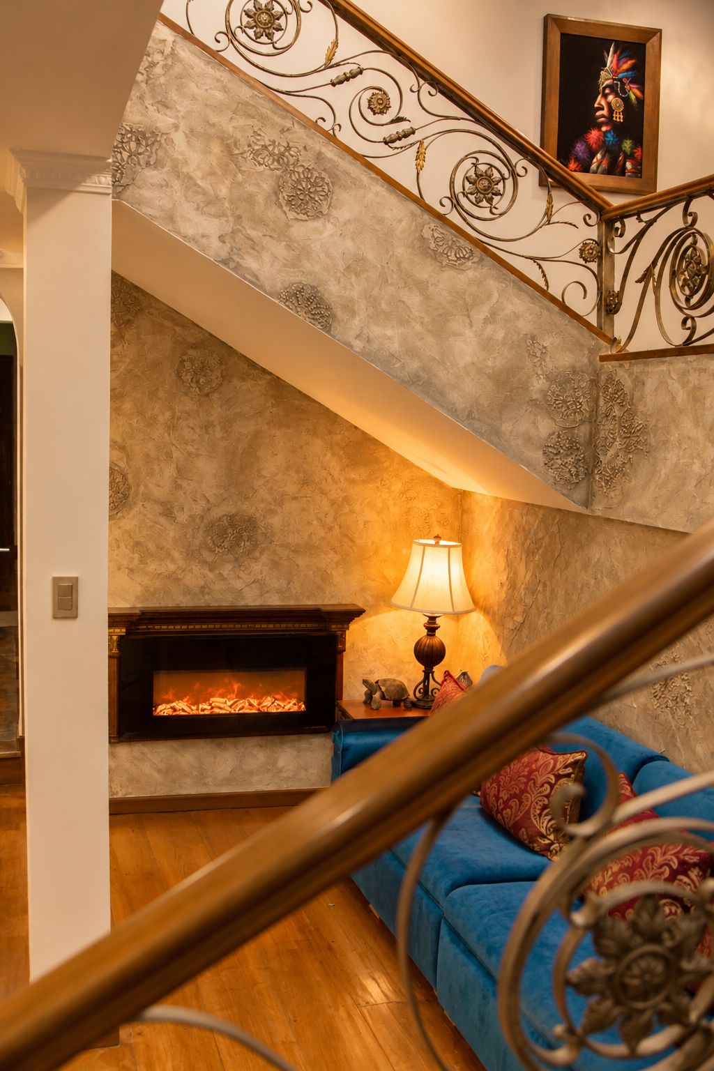 Una escalera con barandillas de metal ornamentadas que conduce a una sala de estar con chimenea, un sofá azul y una lámpara.
