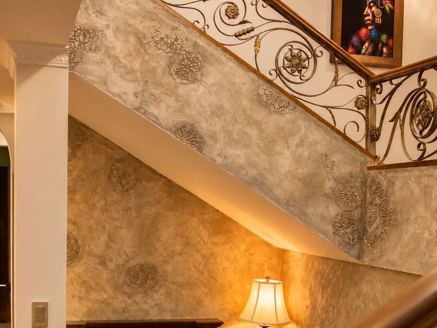 Una escalera con barandillas de metal ornamentadas que conduce a una sala de estar con chimenea, un sofá azul y una lámpara.