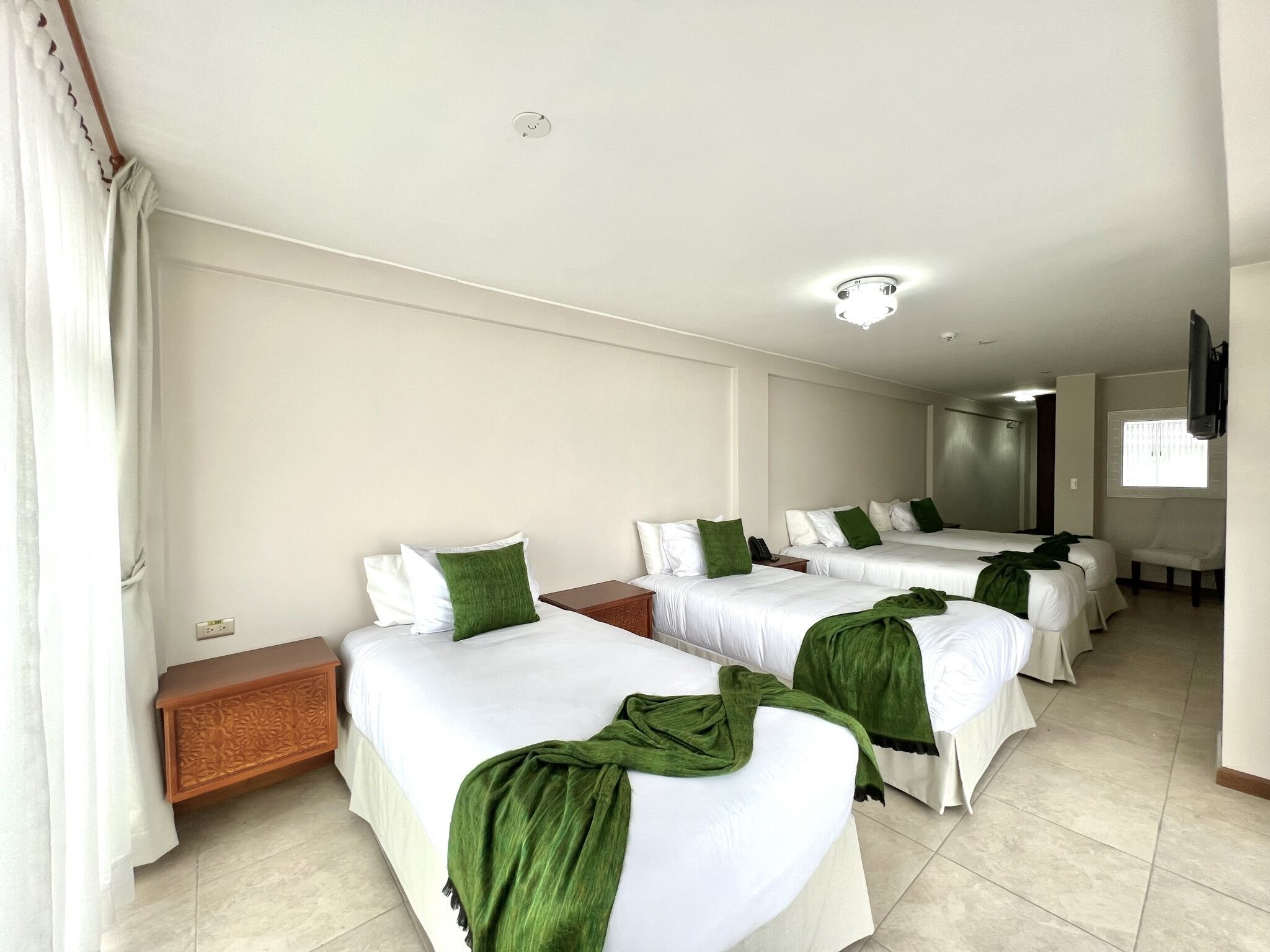 A hotel room with three single beds, each with white bedding and green throw pillows. There is a wooden nightstand, a small table, and a chair. The room has a tiled floor, a ceiling light, and a window with blinds.